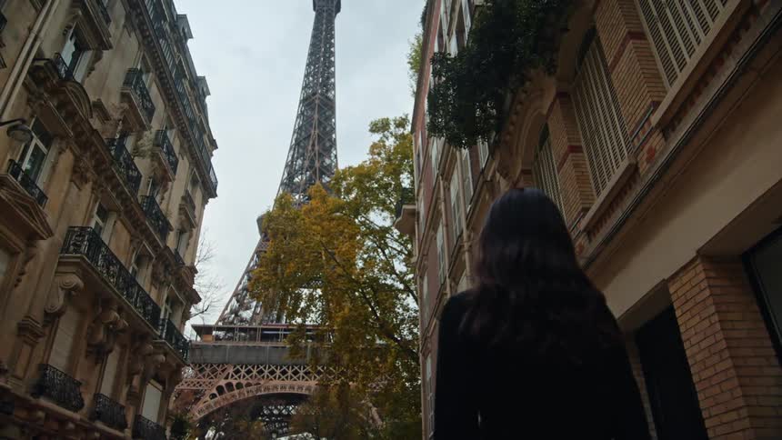 A woman is walking on a street in front of buildings looking up at the eiffel tower