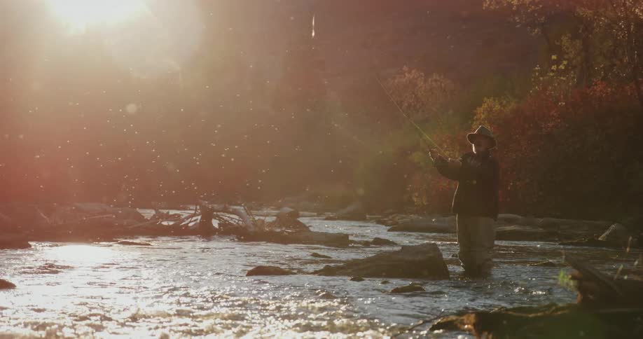 Elderly man with hat beard black coat and gray pants casts a fishing line in the stream as the sun shines brightly