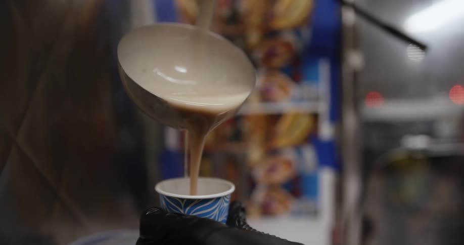 A hand holding a small paper cup as hot liquid batter is poured into it from a ladle