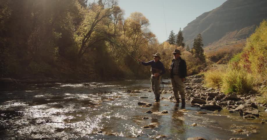 Two men wearing hats are fishing in a stream near the woods and there is a mountain in the distance
