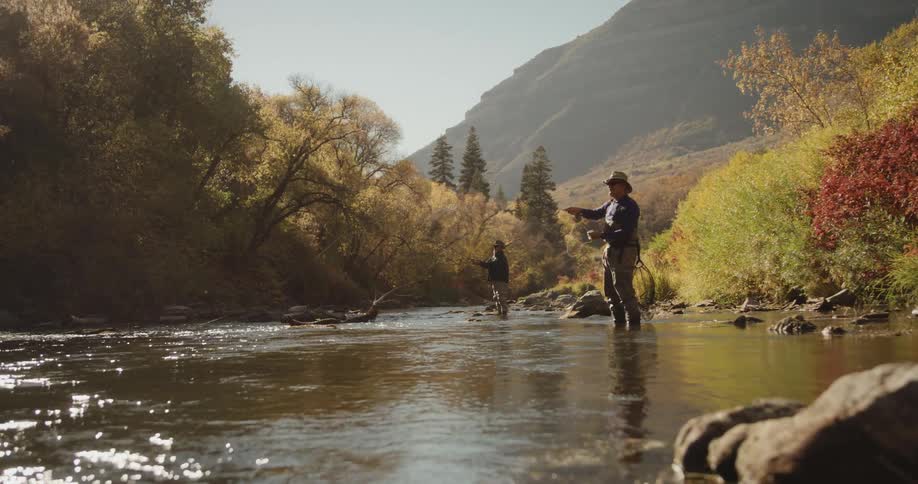 Men wearing hats sunglasses sweater and gray pants cast fishing rods in the stream near the brown trees