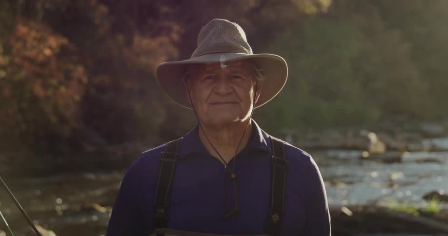 Man wearing a hat stands in front of a river with a fishing rod at morning