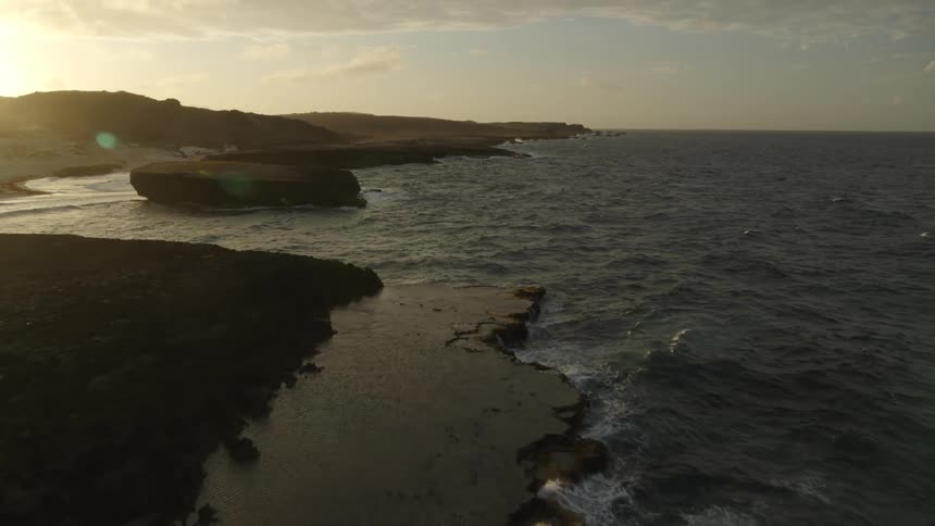 Waves wash ashore the rocky coastline of a small island as the sun sets in the distance