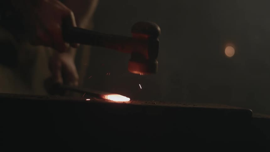 A blacksmith is striking a piece of molten hot metal with a hammer as it glows in the darkness