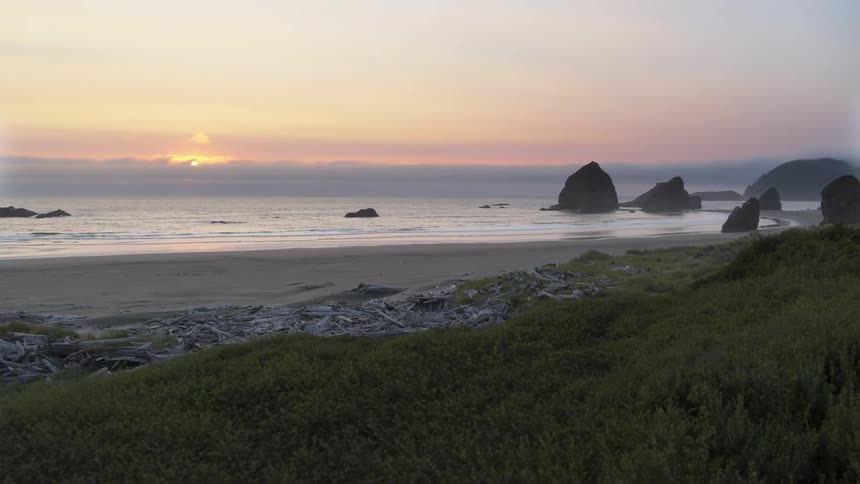 Skimming over lush grassy dunes to reveal sea stacks along a beautiful beach as the sun sets in the distance