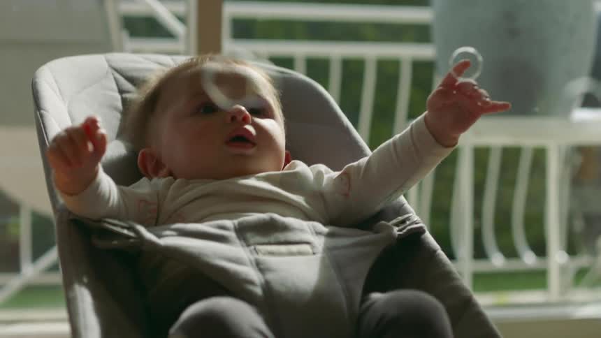 A baby in a rocking carrier reaches out to try and touch bubbles as they fall over him