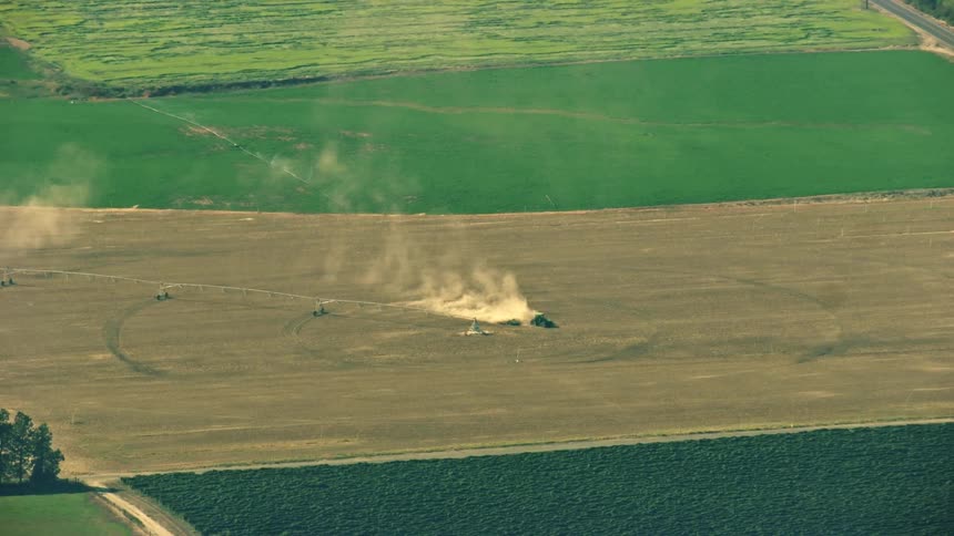 A farming combine John Deere is seen from afar as it drives over a dirt covered patch of land kicking up massive plumes of dust