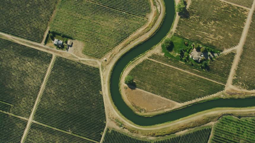 High energy look down and rotating shot of an irrigation canal as it runs through a arid rural areas with orchards and row crops in the otherwise dry desert