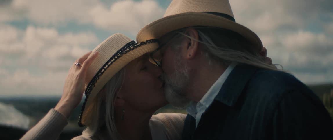 A man and a woman wearing matching hats are standing outside overlooking a beach as they kiss