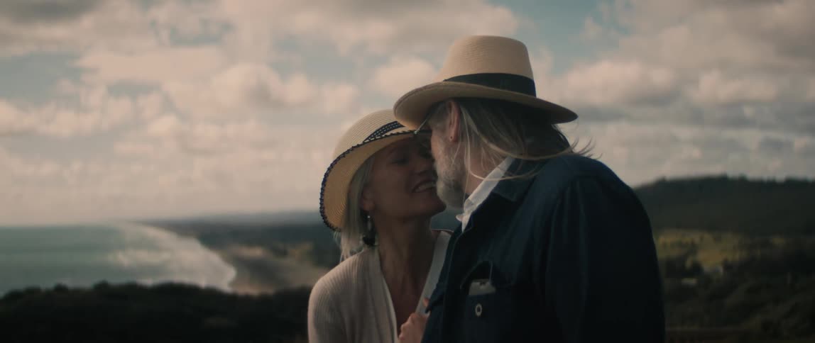 A man and a woman wearing matching hats are standing outside overlooking a beach as they kiss