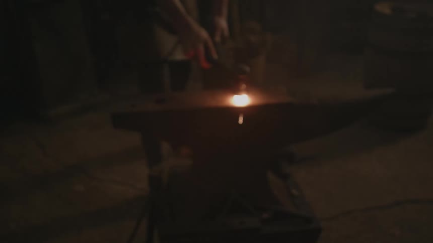 A blacksmith heats a piece of metal on flaming coals and takes it to his anvil where he hits it with a hammer as sparks fly and it glows in the darkness