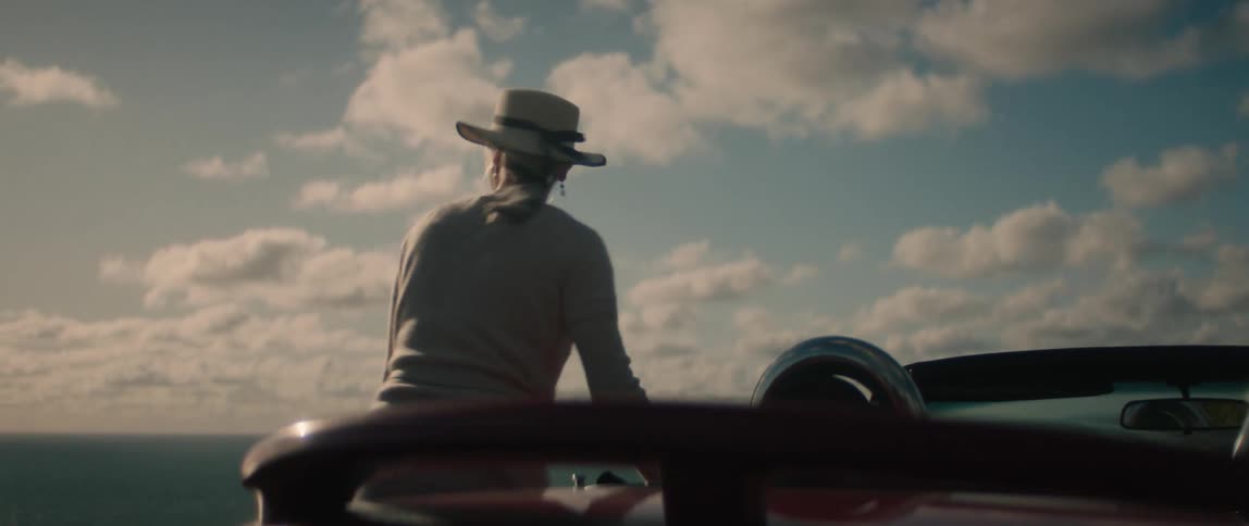 A woman holds onto her hat while stepping out of her convertible parked along the coast