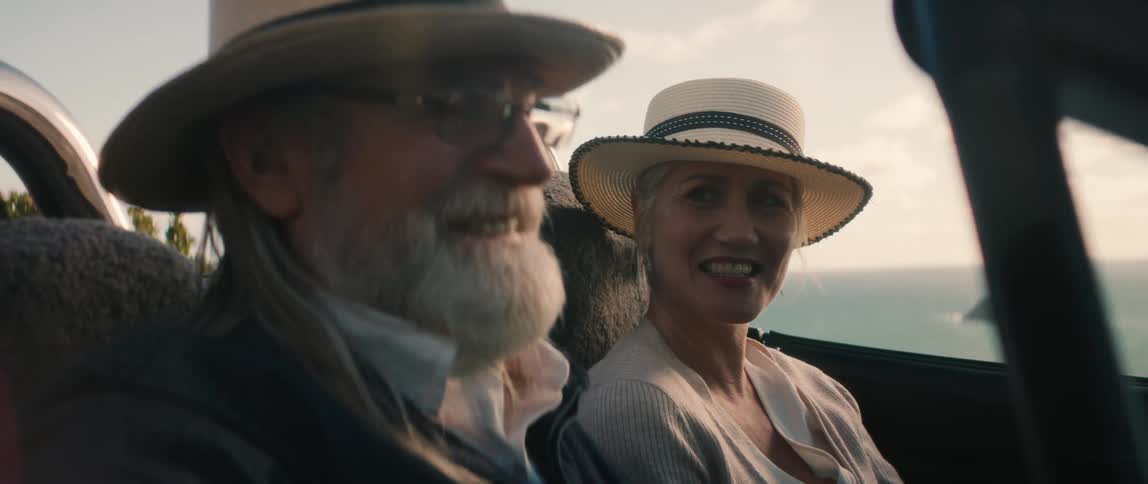 Elderly man with hat glasses and beard sits in convertible car next to woman wearing hat earrings and white shirt laughing and talking