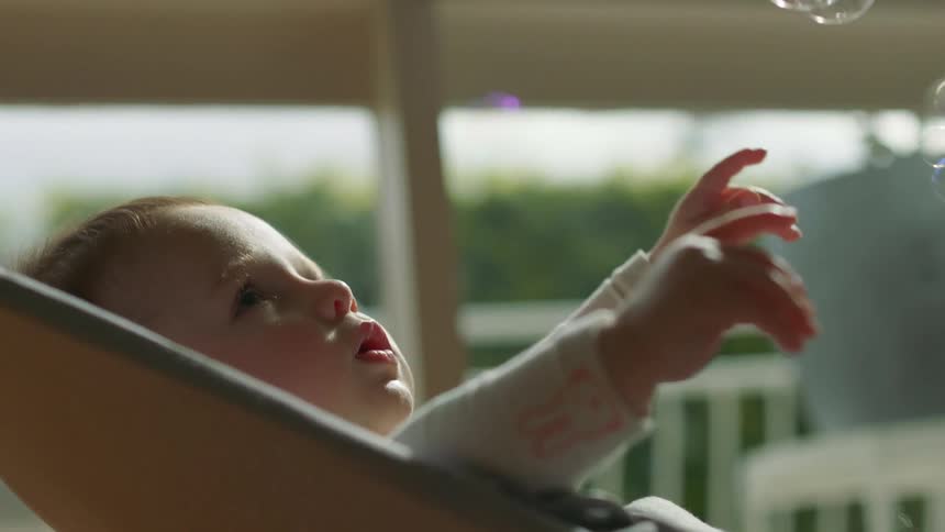 A baby lays back in a chair while watching bubbles fall from above