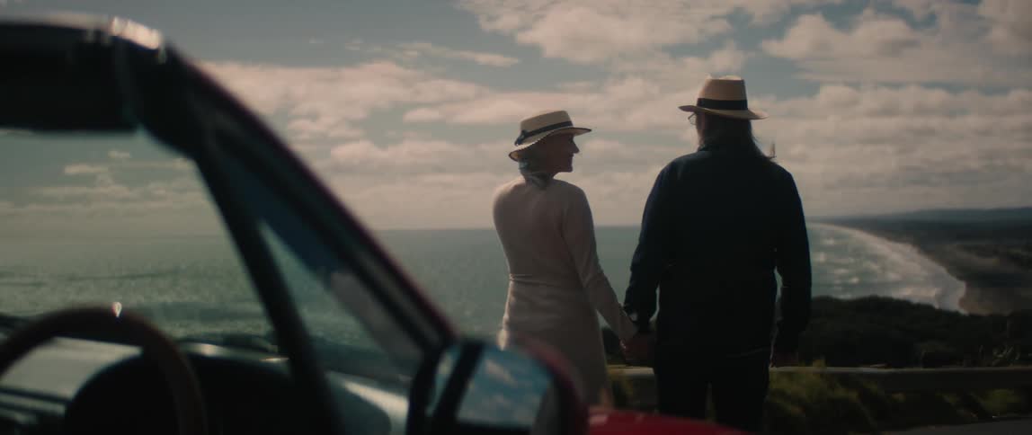 Man and woman stand in front of a car as they hold hands and look out at the ocean