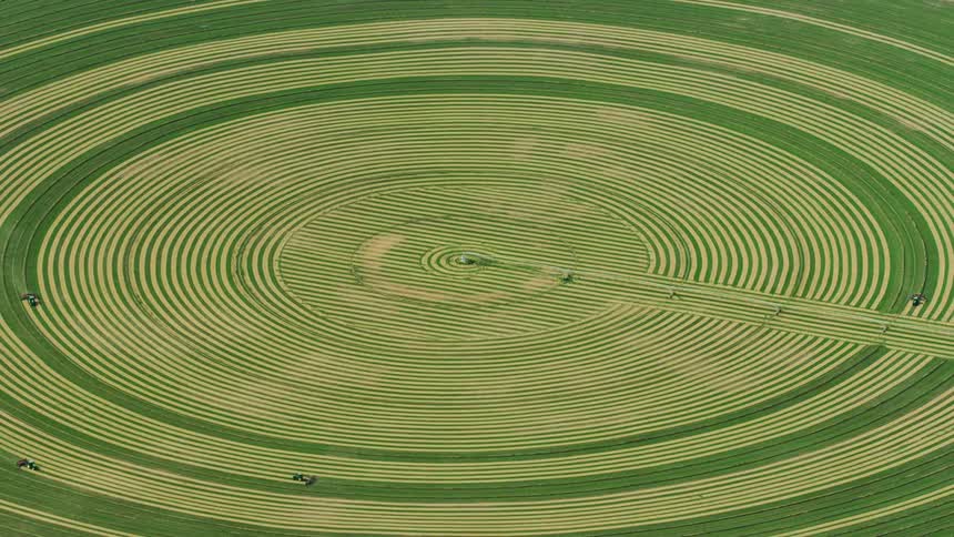 Slow zoom out reveal in this high altitude aerial of combines harvesting alfalfa showing perfectly symmetrical circular field with center pivot irrigation