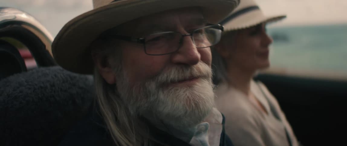 An elderly man with glasses and a beard is smiling as he drives along the coast with a woman