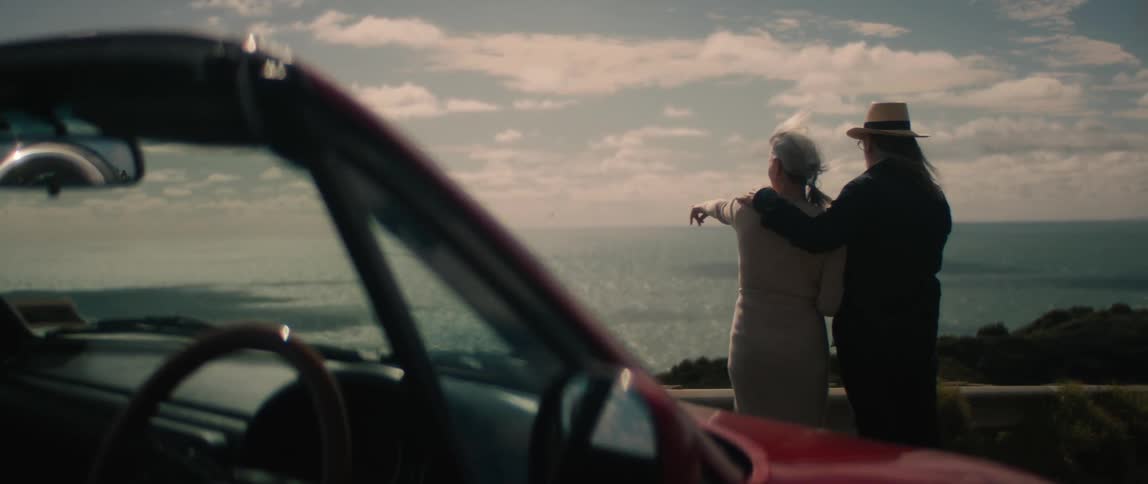 A man and a woman are standing along the coast near their automobile looking at the waves rolling in and enjoying the outdoors
