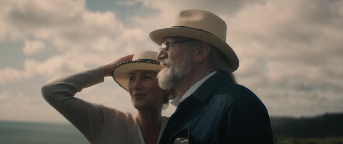 A man and a woman wearing matching hats are standing outside overlooking a beach as they talk and she has her hand on her hat