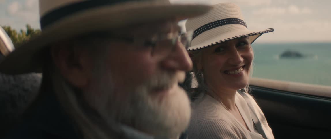 A man and a woman are sitting inside a convertible automobile overlooking the water smiling and laughing