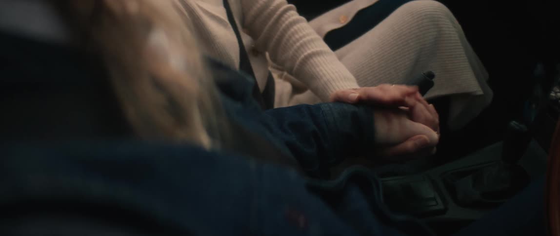 Woman sits next to man with long hair and dark blue jacket holding hands in convertible car