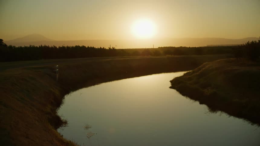 Sunset over an irrigation canal through a rural area with silhouettes of trees and mountains in the distance under a sunset sky