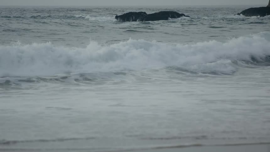 Waves crash onto a beach and over some rocks in the background