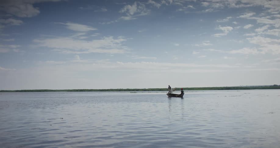 One man casts fishing net while standing in boat as another person paddles the boat