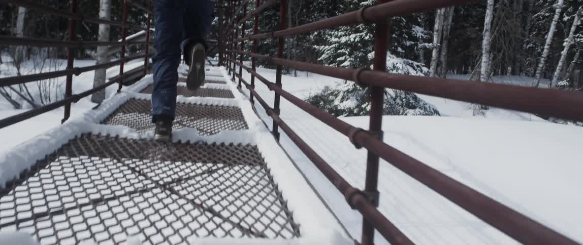 Woman wearing brown shoes jeans and coat runs across rusty iron bridge in the snowy field