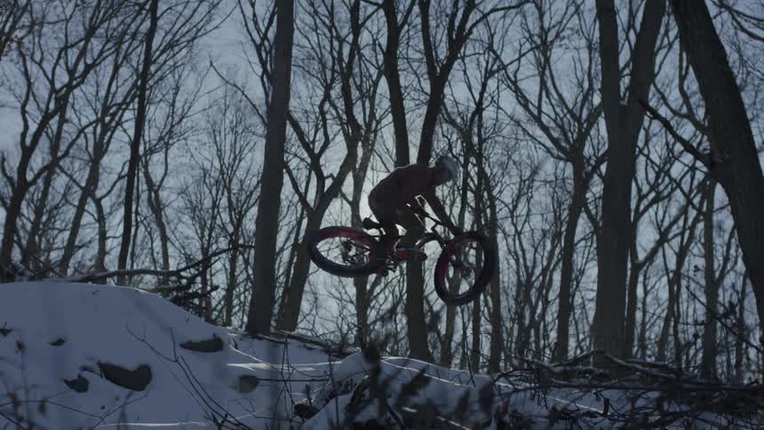A teenage boy rides his bike over a hill in the snow covered woods as the sun shines in a hazy sky through the bare trees