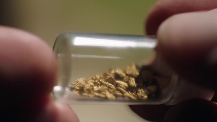 A man is holding a small jar containing gold granules in his fingers and he is turning it so they move