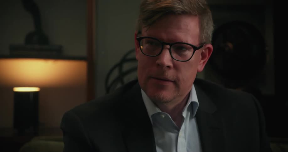 Man wearing glasses blazer jacket and light blue shirt exhales and shakes his head slightly
