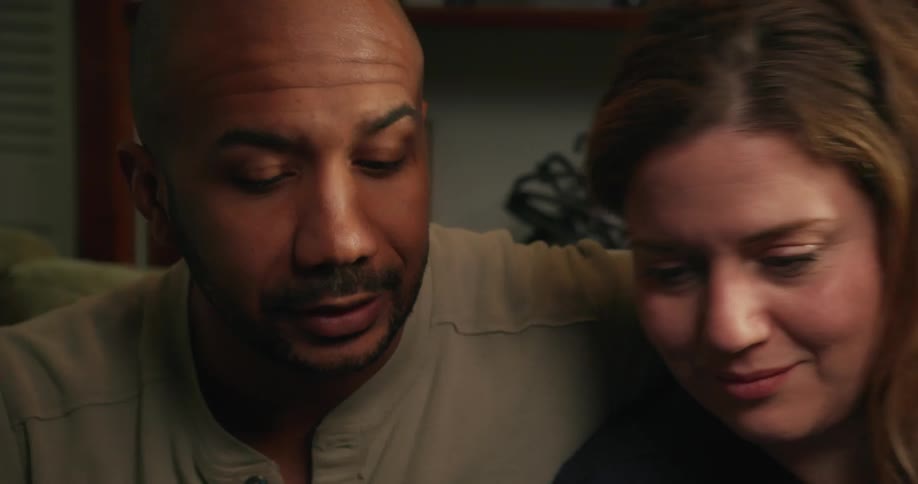 A man is sitting on a couch talking and he puts his arm around the woman sitting next to him and they smile