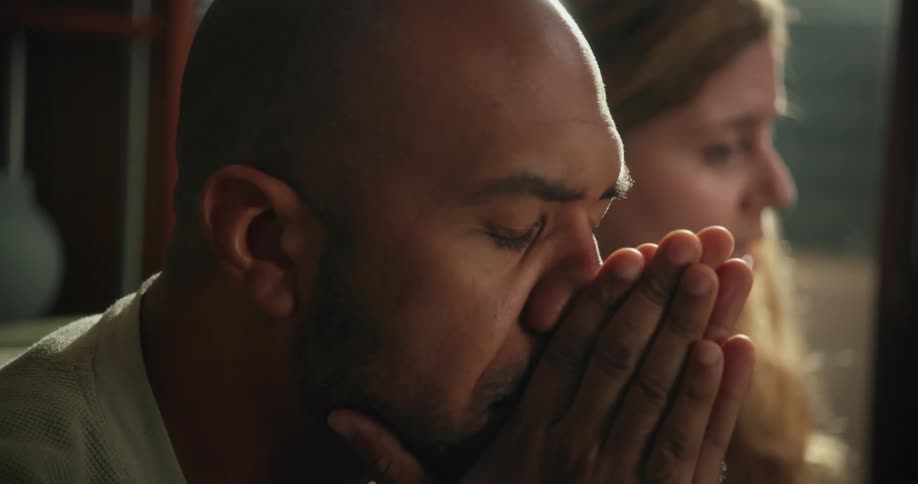 A man and a woman are sitting on the living room couch and as she speaks he puts his hands in front of his face