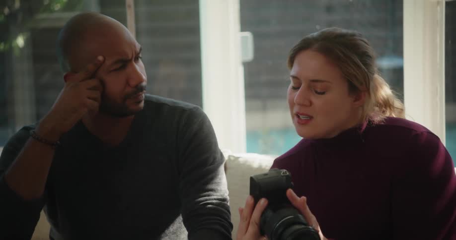 Man and woman sit on couch near glass doors discussing pictures on camera