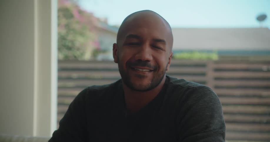 A man is sitting on a couch smiling and scratching his head as he nods and smiles more