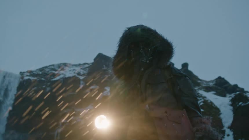 A man wearing a winter coat is standing outside in a snowstorm holding a flashlight and there are mountains in the distance