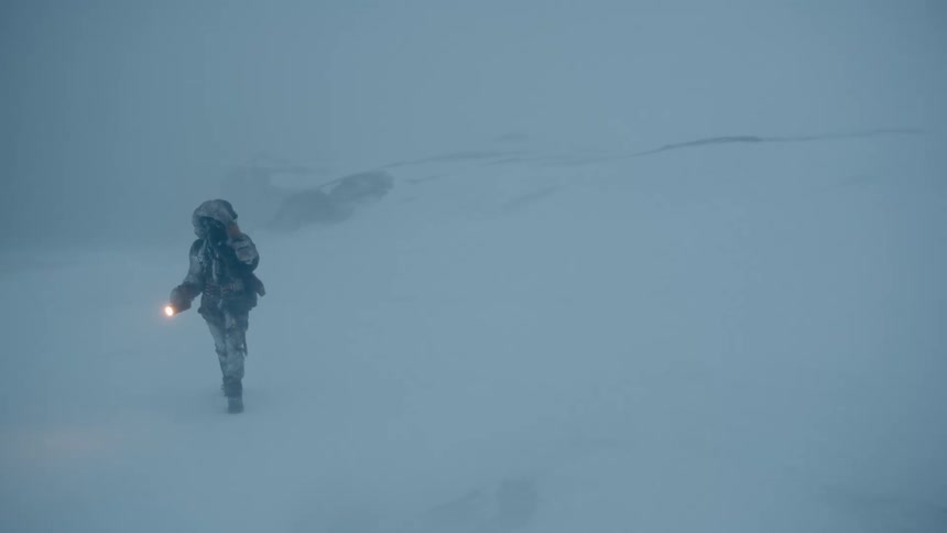 A person carries a flashlight and holds onto their coat hood while walking through a snowstorm