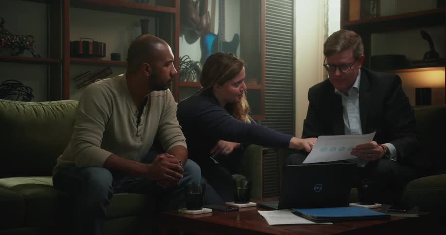 Man wearing glasses and blazer jacket sits on couch holding a piece of paper and couple looks at it discussing the terms in the living room at home