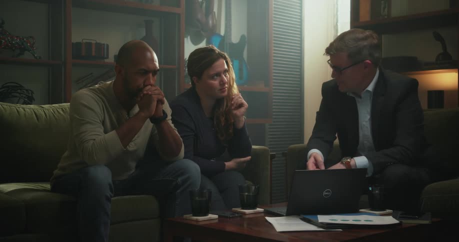 A man speaks to a couple while gesturing at a laptop as they all sit together in the living room of a house