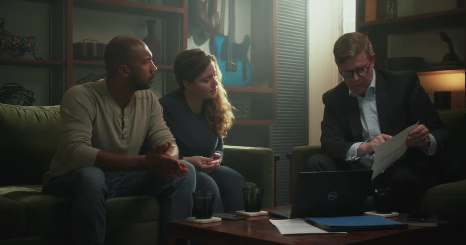 A man in a suit speaks to a couple while showing them a sheet of paper in the living room of a house during the day