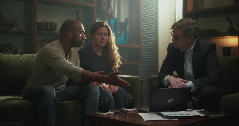 Couple argues with each other sitting on couch near man wearing glasses and blazer jacket checking his laptop in living room at home