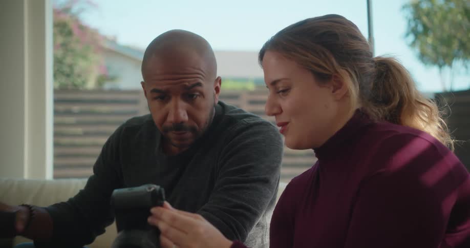 A man and a woman are sitting in the living room and she is holding a camera and showing him pictures as he takes a drink from his glass