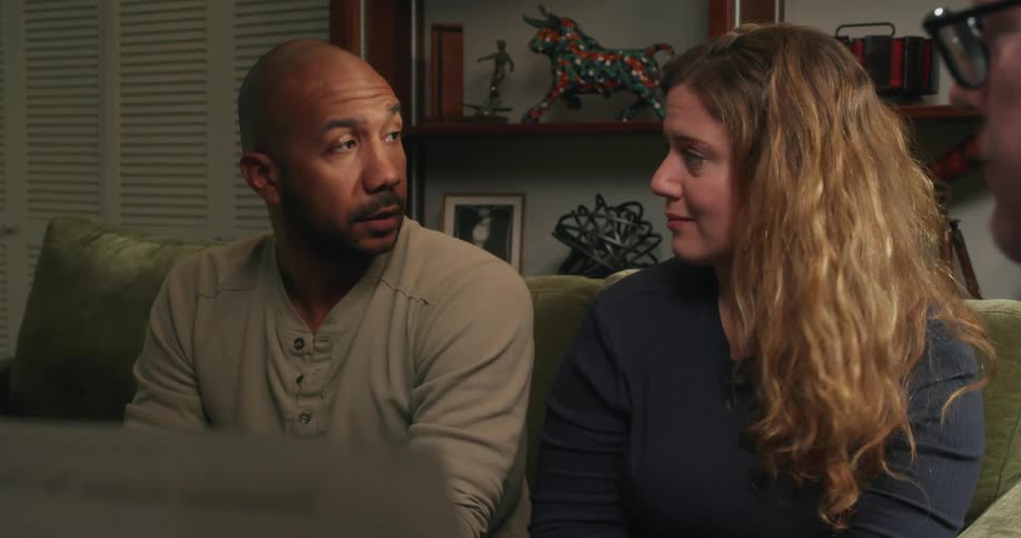 Three people are sitting in a living room talking and a man is holding a piece of paper and the other man shakes his hand and puts his arm around the woman sitting next to him
