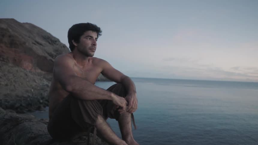 A shirtless man sicks on a rock near the ocean at dusk