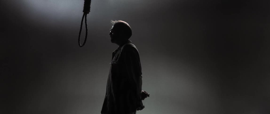 A man with hands tied behind his back looks up toward a hangman's noose