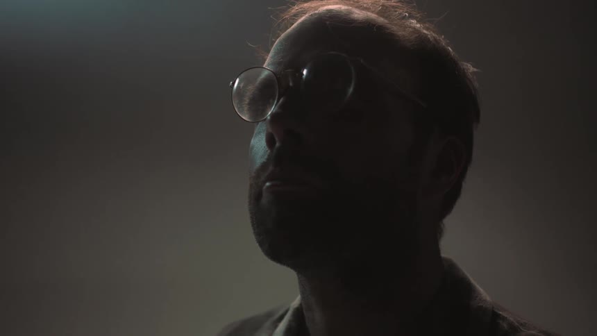 A man wearing glasses looks upward in a dim room