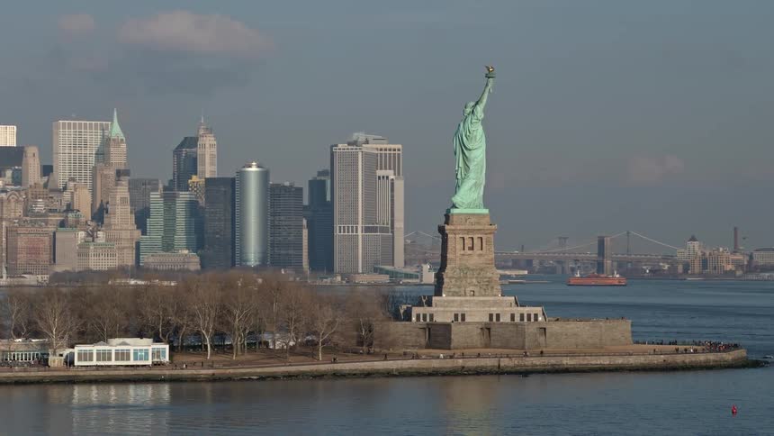 Aerial shot of the Statue of Liberty orbiting around its torch side and revealing the Lower Manhattan skyline in the background during winter
