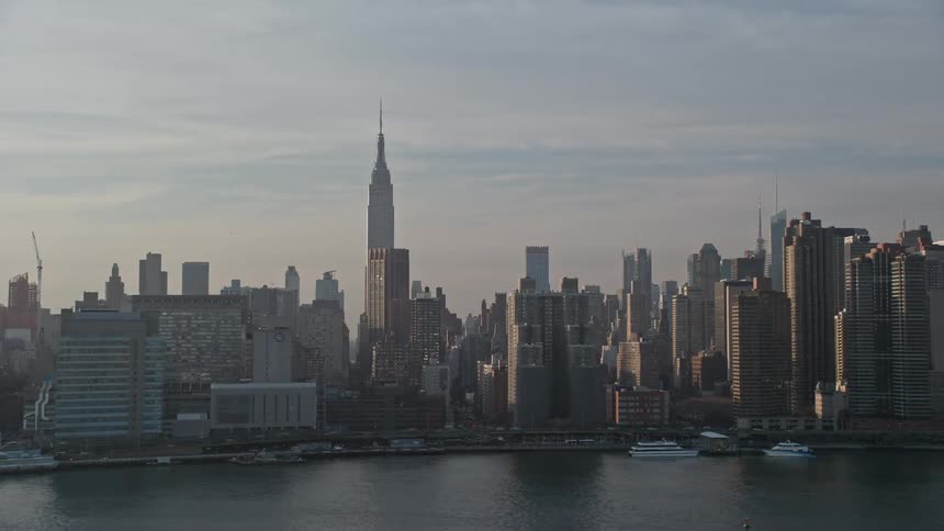 Aerial view of Empire State Building and Manhattan condos on the East River waterfront with boats docked and cars traveling under a cloudy sky