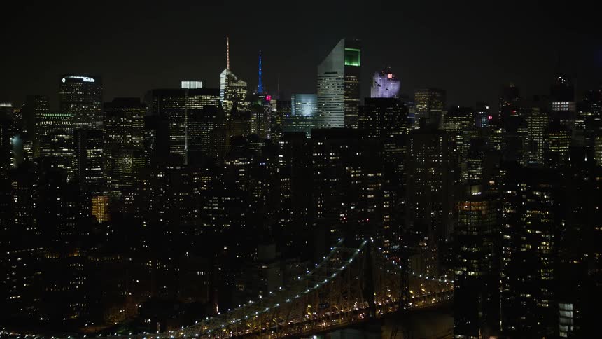 Aerial footage reveals Midtown Manhattan skyscrapers as vehicles travel on the bridge over the East River next to the illuminated city buildings at night during winter season